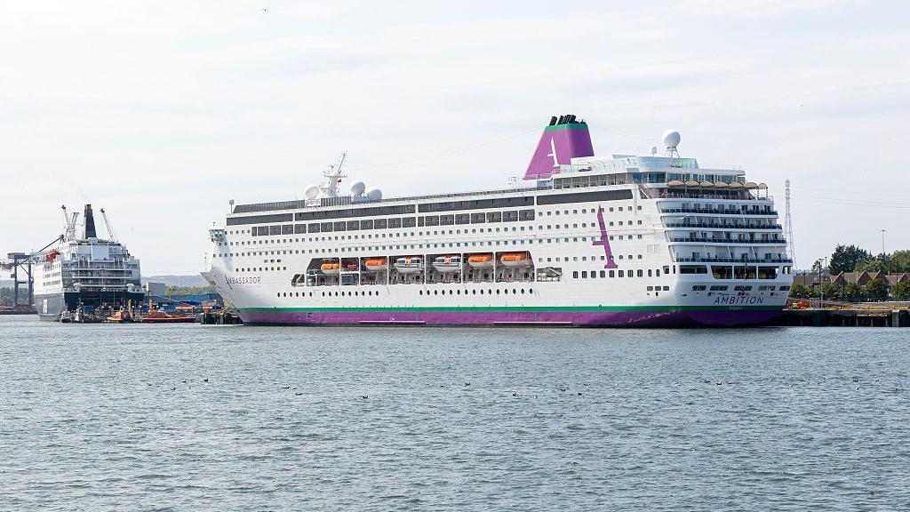 Imagen de un crucero en el mar con pasajeros en la cubierta, algunos de ellos con mascarillas
