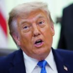 Donald Trump hablando en un podio con banderas de Estados Unidos detrás, sonriendo tras anuncio de suspensión de ataques a Irán